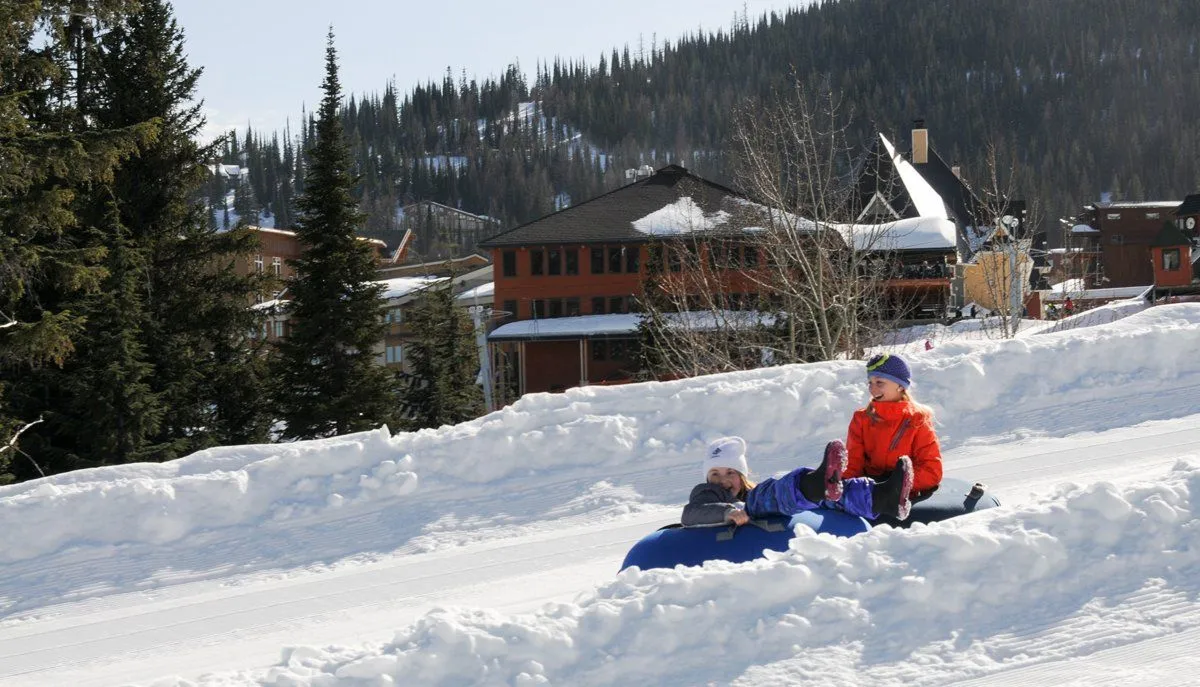 Two people sledding down a snowy hill near a lodge in a mountain setting