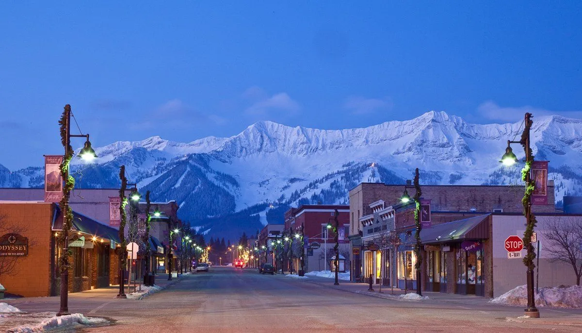 Snowy small-town street at dusk with lit storefronts and mountains in the background