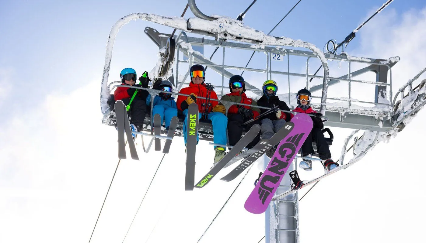 Skiers in colorful gear ride a chairlift with skis and a snowboard against a snowy sky.