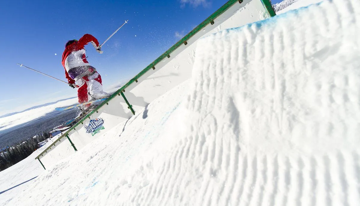 Skier in red jacket airborne over a green rail on a snowy slope under a blue sky