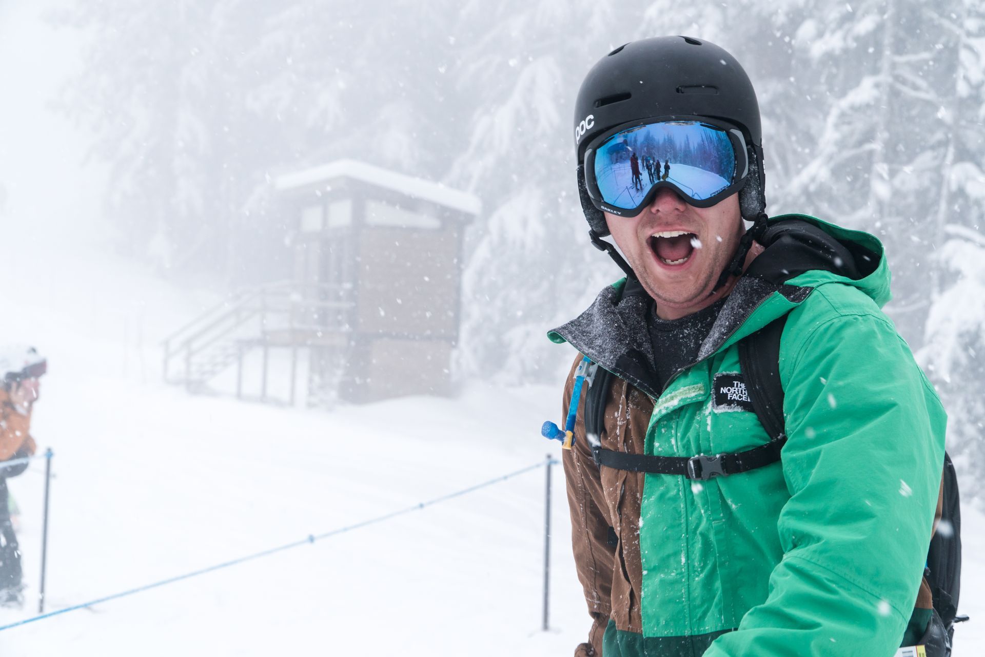 Smiling skier in green jacket and blue goggles at a snowy mountain lift