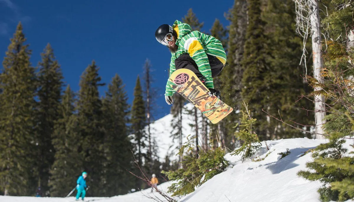 Snowboarder in green jacket midair over a snowy slope with pine trees under a blue sky