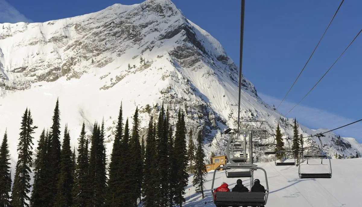 Chairlift ascending a snowy mountain with pine trees under a clear blue sky