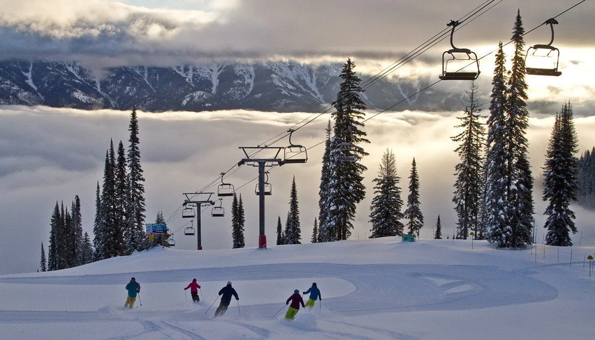 Skiers on a snowy slope beneath chairlifts and tall evergreens, with mountains in the distance.
