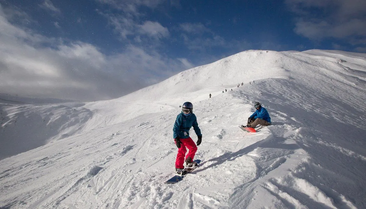 Two skiers descend a snowy mountain ridge under a cloudy sky.