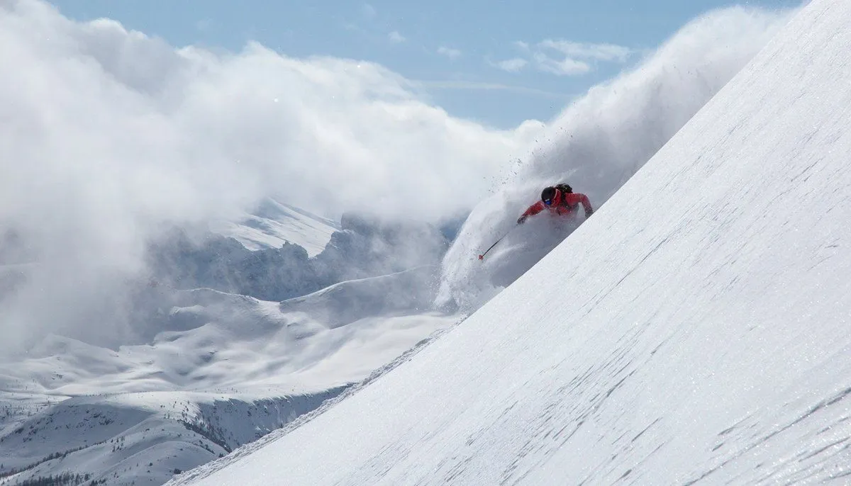 Skier in red carving down a snowy mountain slope amid clouds