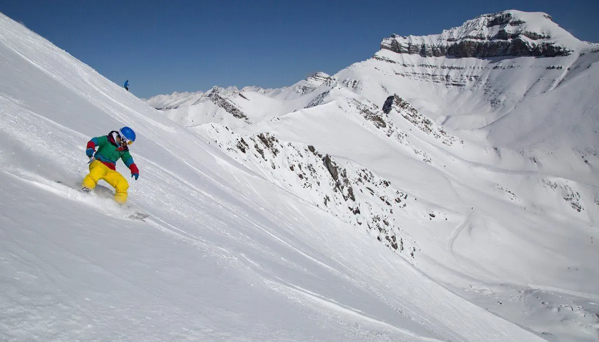 Skier in colorful gear carving down a steep snowy mountain slope under a blue sky