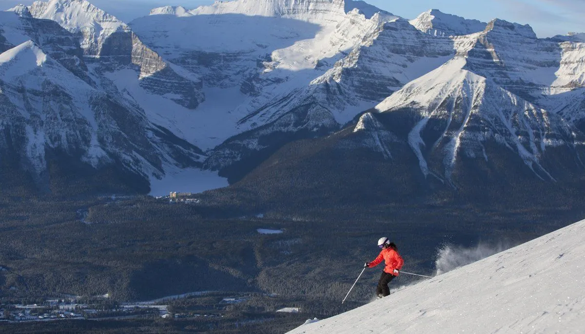 Skier in a red jacket descending a snowy slope with a mountain range in the background