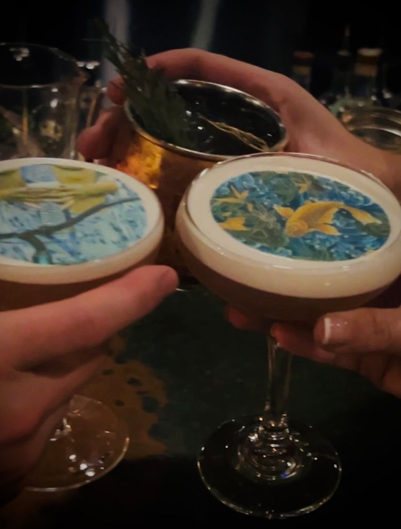 Two hands clinking cocktails topped with printed circular garnishes in a dim bar setting