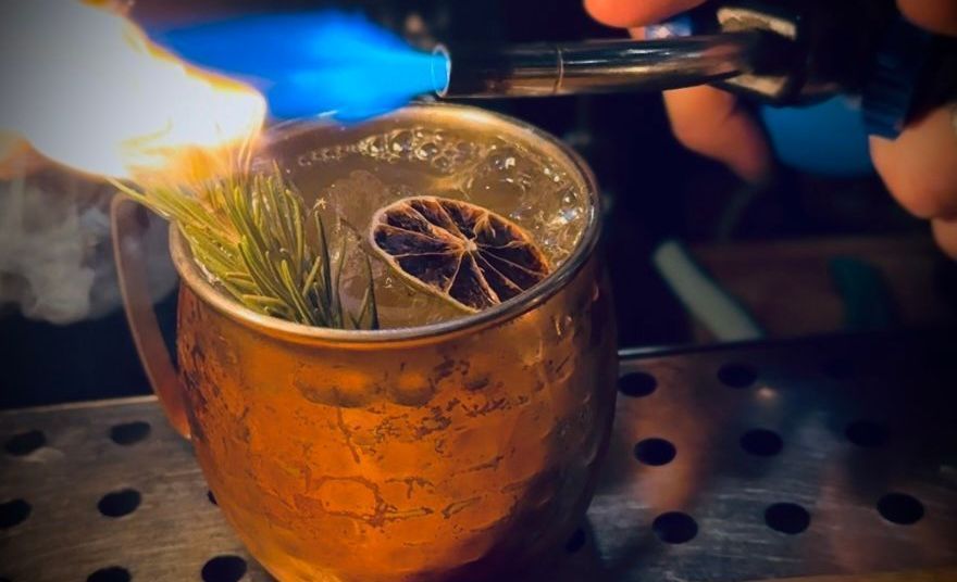 Cocktail being flame-garnished with a dried citrus slice and herbs on a bar counter