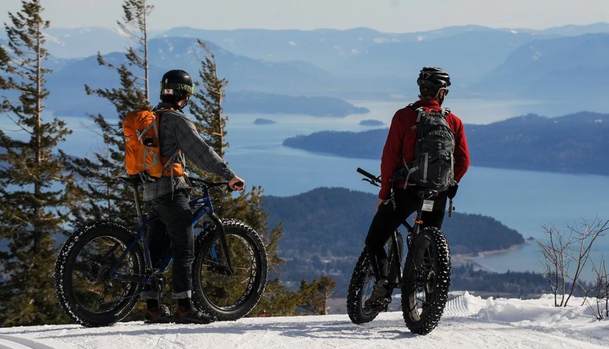 Two cyclists with fat bikes overlook a snowy mountain lake and distant blue hills