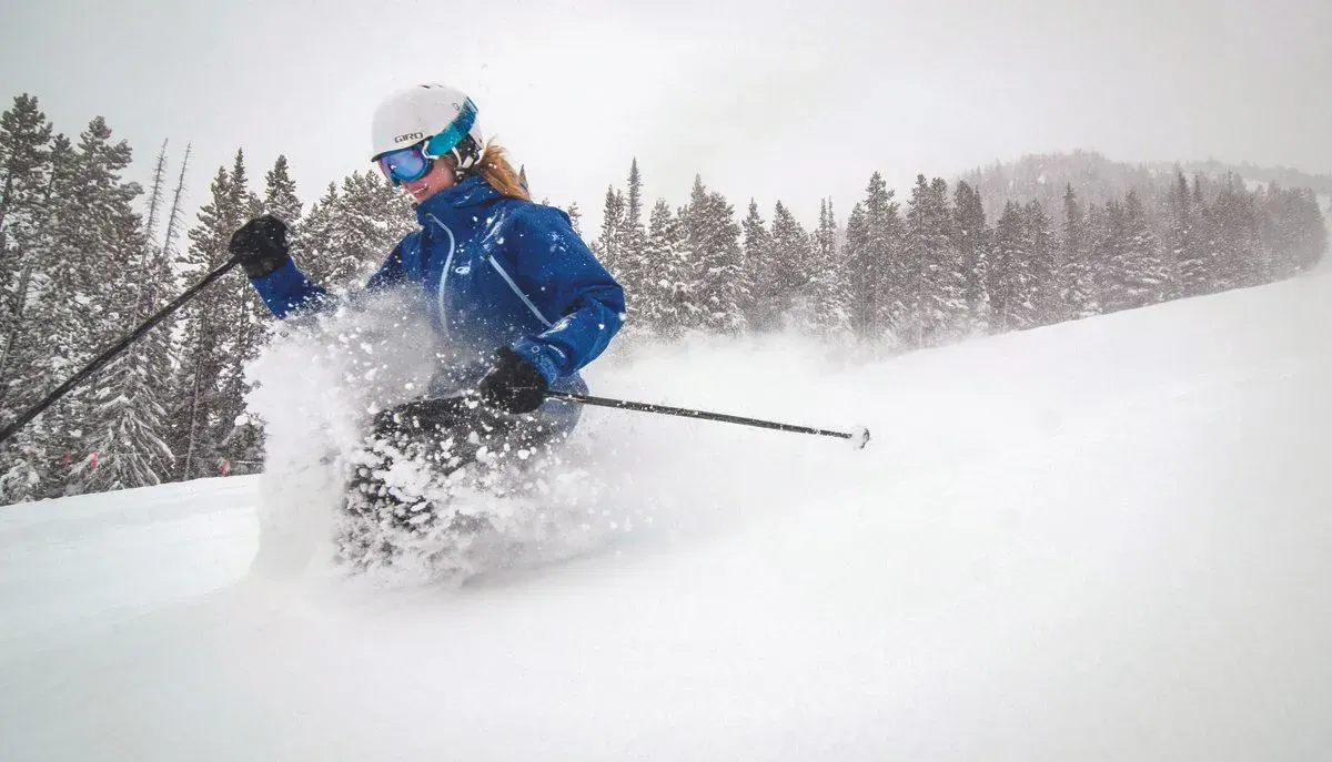 Skier in blue jacket carving through deep powder on a snowy slope, with trees in the background.