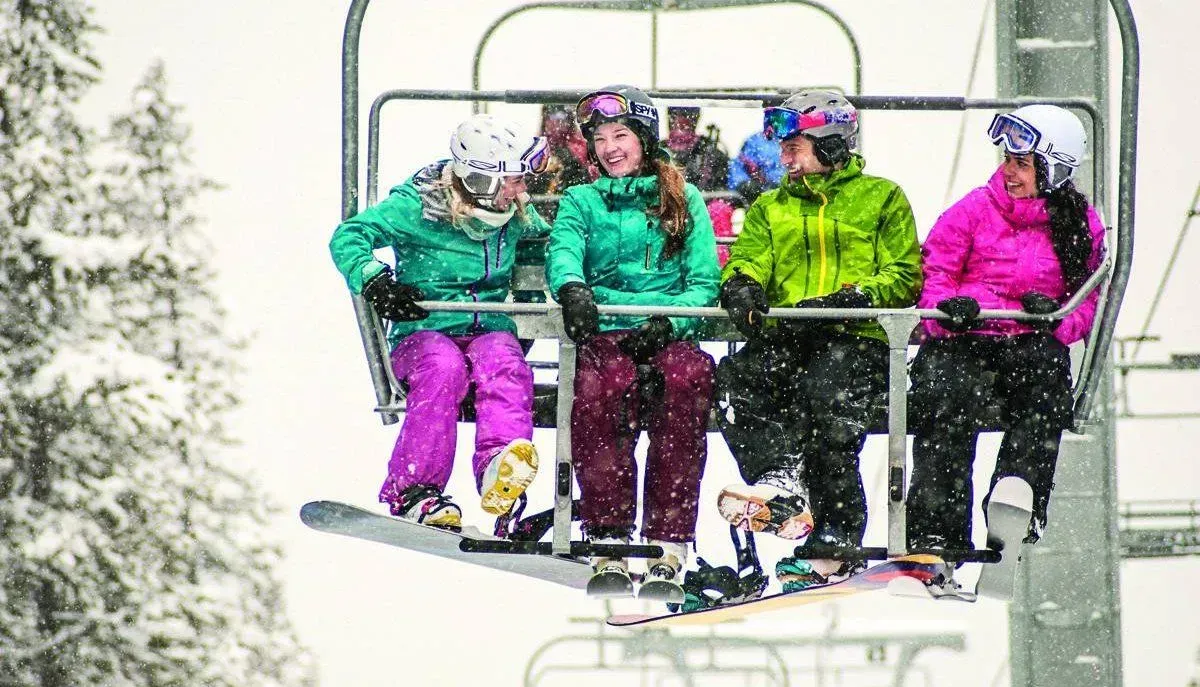 Four skiers in bright jackets ride a chairlift through a snowy mountain scene.