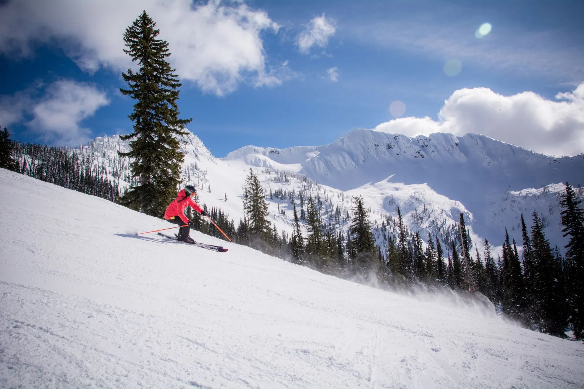 Skier in red carving down a snowy slope with mountains and pine trees in the background