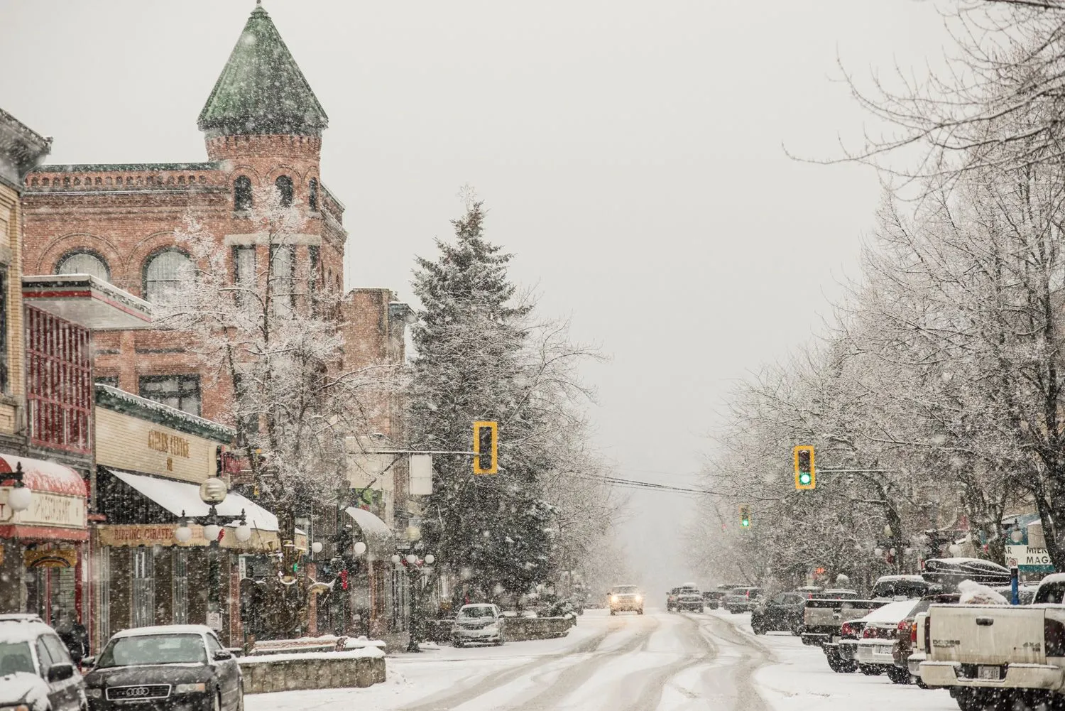 Snowy small-town street with historic brick buildings, parked cars, and trees in winter.
