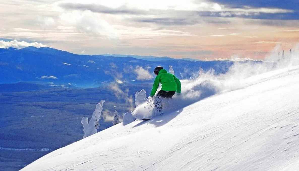 Skier in a green jacket carving down a snowy mountain slope with distant peaks and clouds