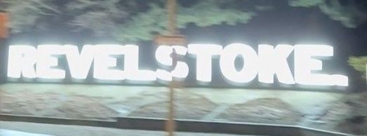 White neon “REVELSTOKE” sign on a dark background with trees behind it.