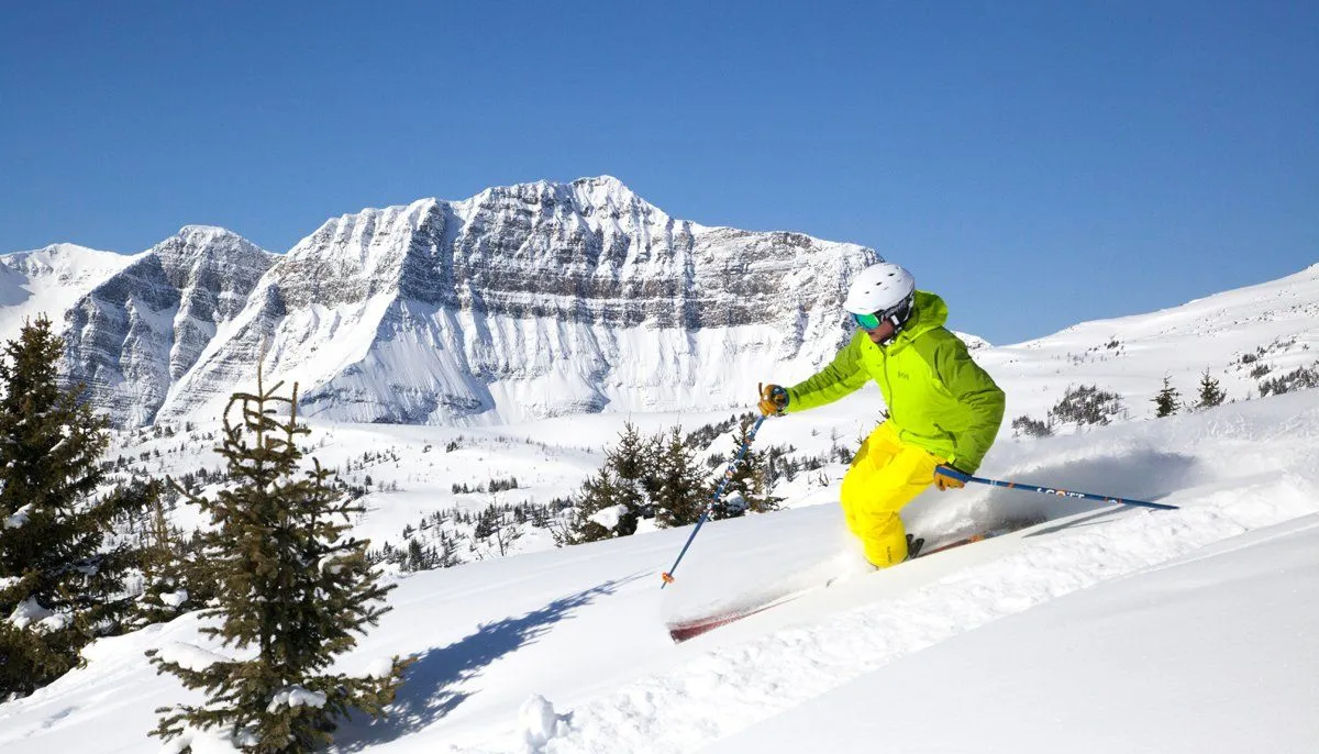 Skier in bright yellow jacket carving down a snowy slope with rugged mountains under a clear blue sky
