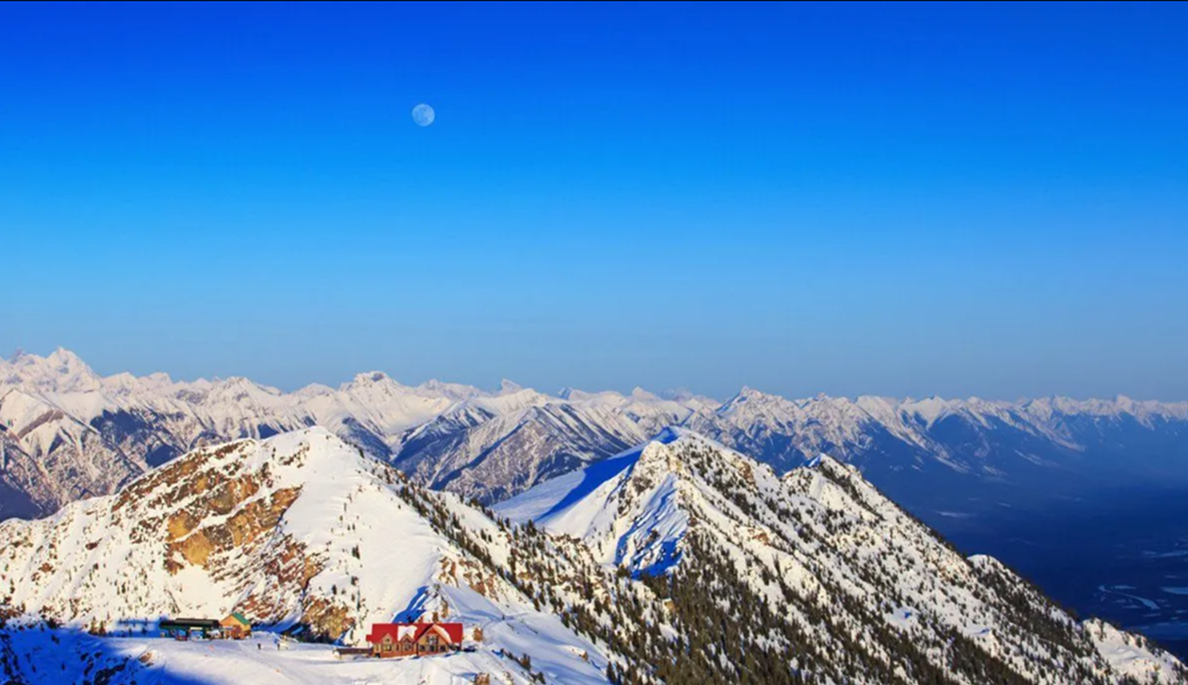 Snowy mountain peaks under a clear blue sky with the moon visible overhead