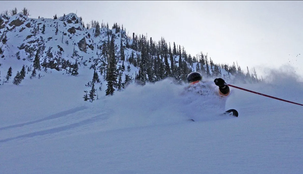 Snowy mountain slope with two skiers descending beneath a rocky ridge and pine trees