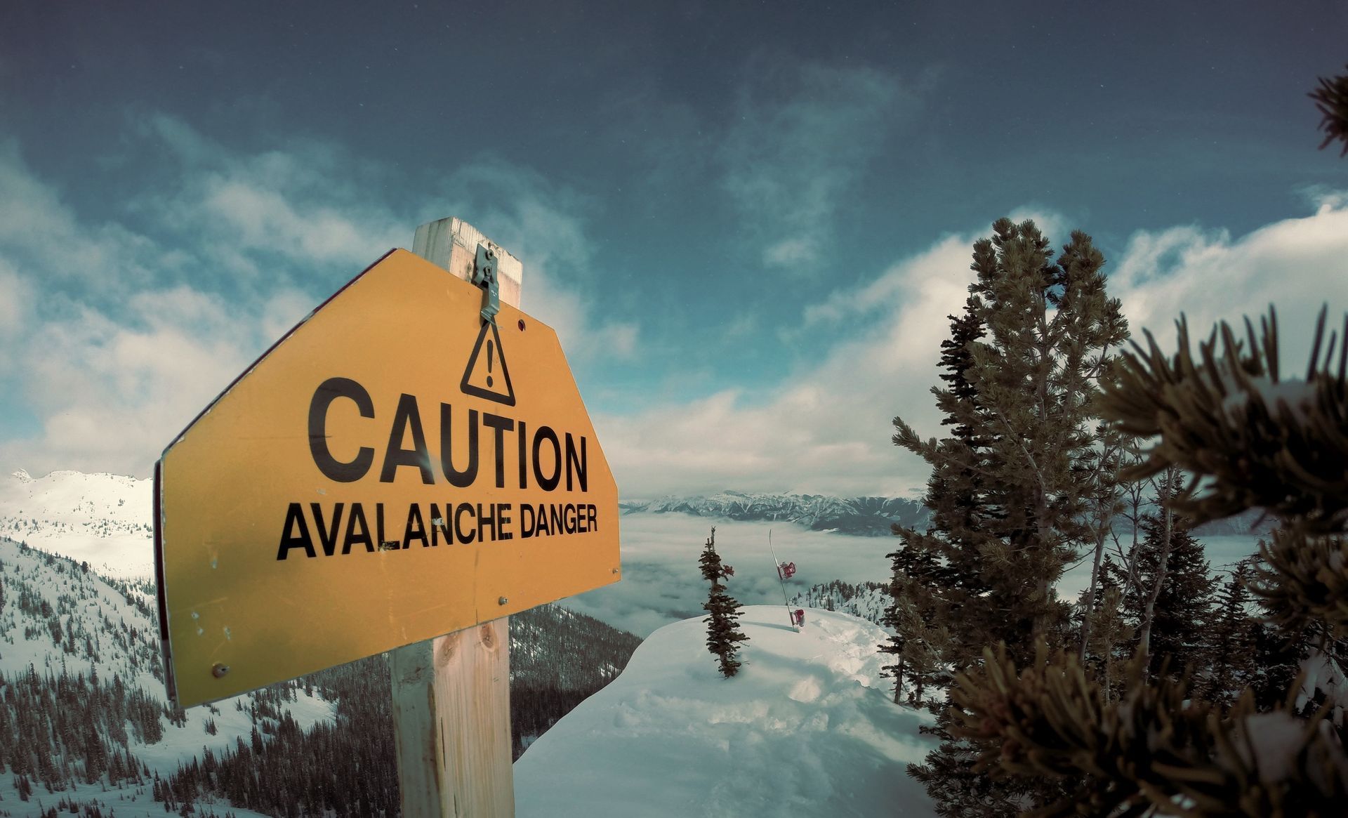 Yellow avalanche caution sign by a snowy mountain trail with trees and cloudy sky.