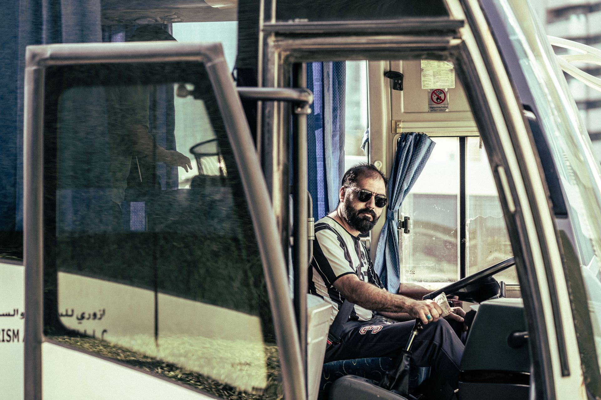 Bus driver seated at the wheel, looking out through the front door of a parked bus.