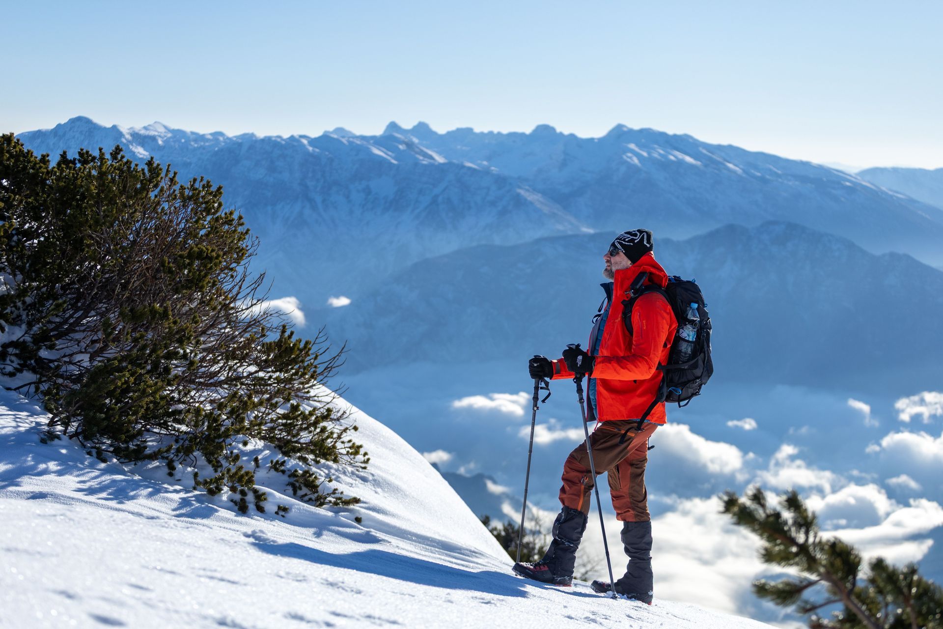Hiker in red jacket stands on snowy mountain ridge with trekking poles, overlooking distant peaks and clouds