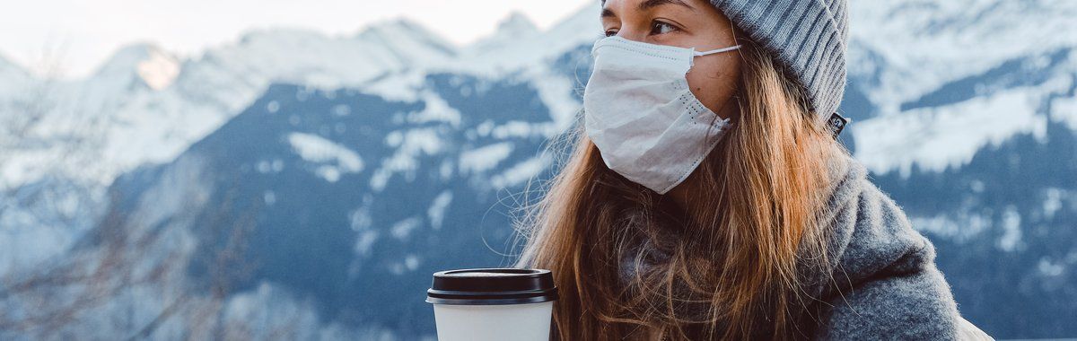 Woman in a gray beanie and face mask holding a coffee cup with snowy mountains behind her