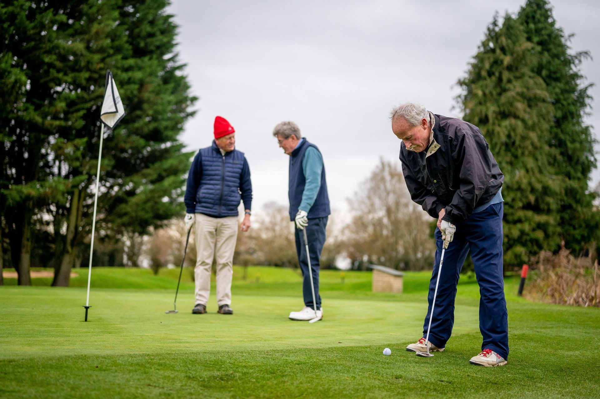 Three people putting on a golf green, with a flagstick and trees in the background.