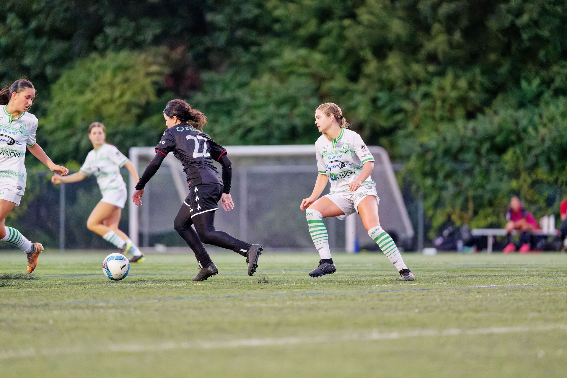 Soccer players in green and black uniforms chase the ball on a grassy field.