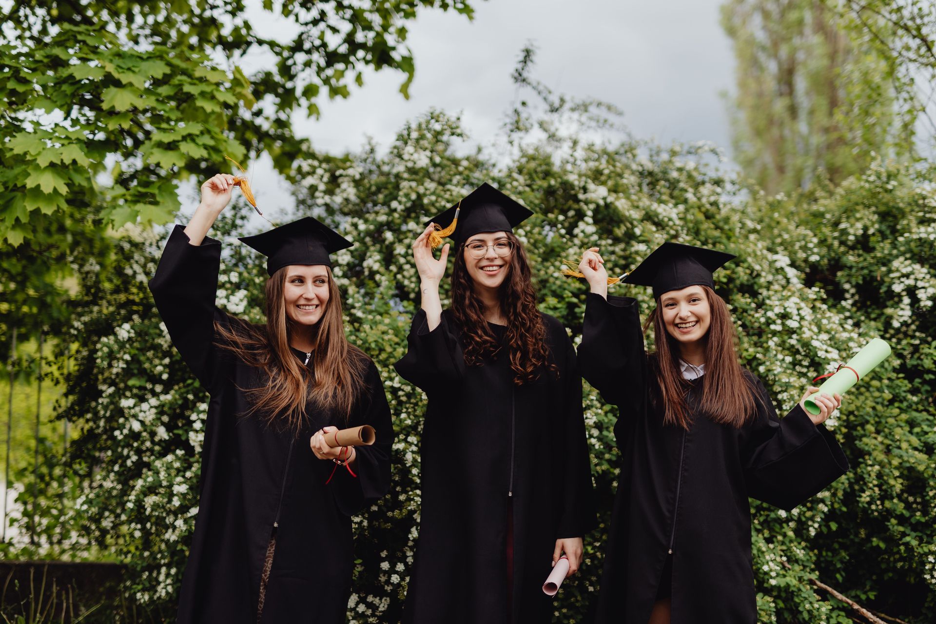 Three graduates in black gowns and caps celebrate outdoors, holding diplomas and raising their hands.