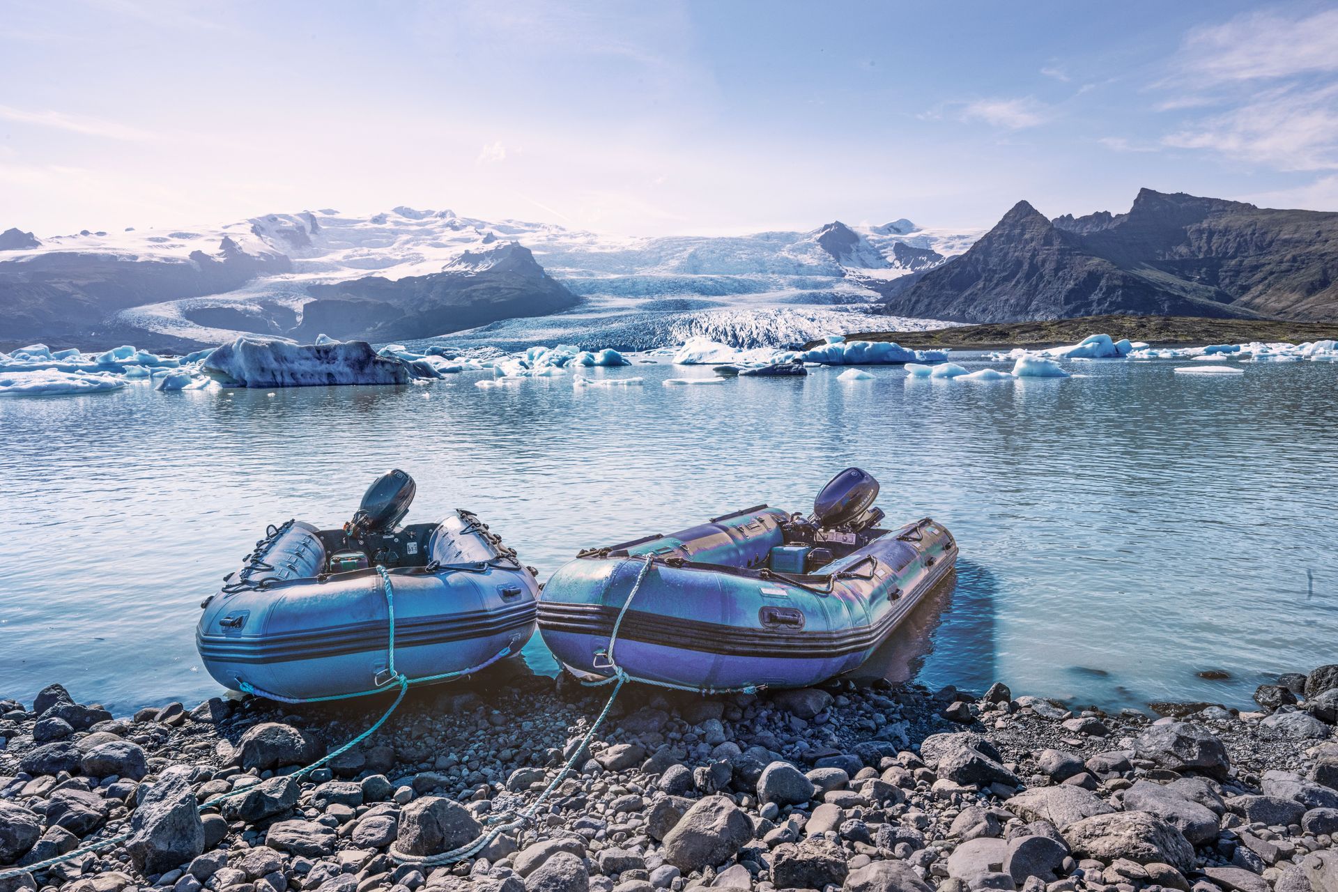 Two small blue boats on a rocky shore, floating on icy water with glaciers in the distance.