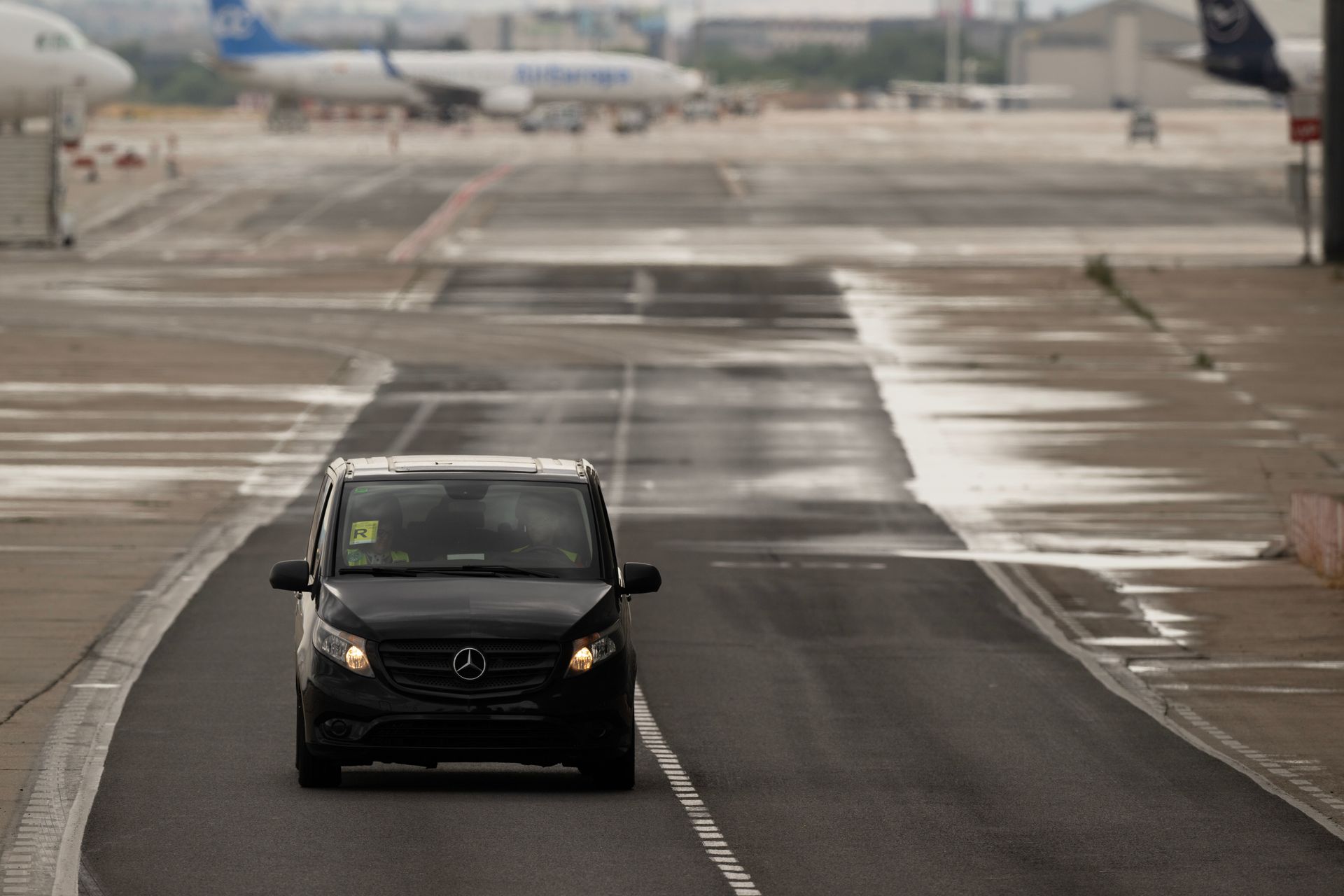 Black car driving on a wet airport runway under overcast skies