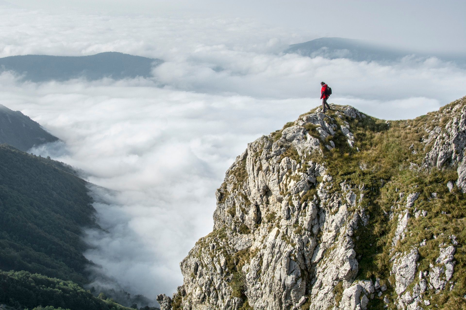 Hiker in red standing on rocky mountain peak above clouds and misty valleys