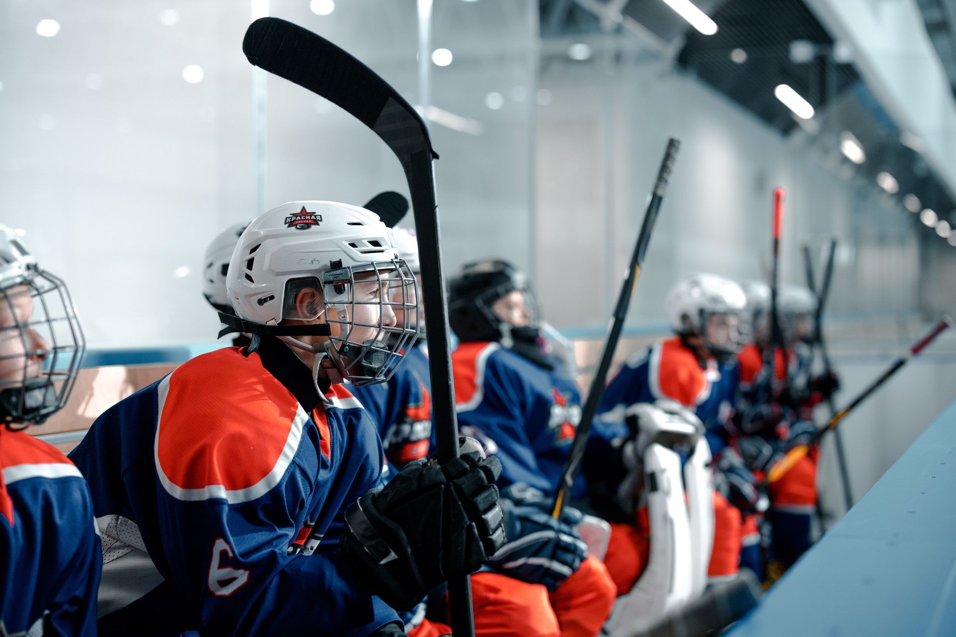 Hockey players in blue and orange uniforms sitting on the bench, holding sticks in an indoor rink