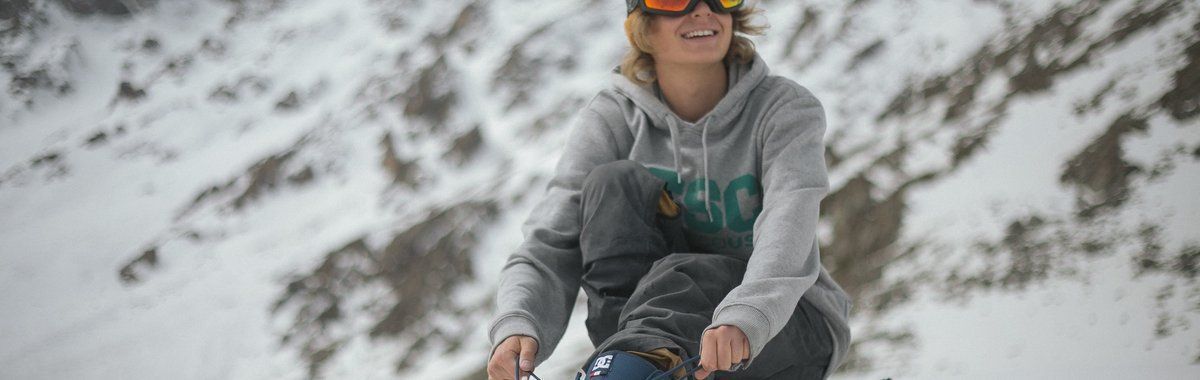 Person in a gray hoodie and orange goggles skiing down a snowy mountain slope.