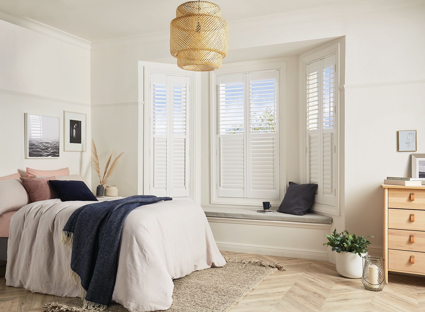 Bedroom with white shutters on bay window, bed with light pink bedding, natural rug, and woven light fixture.