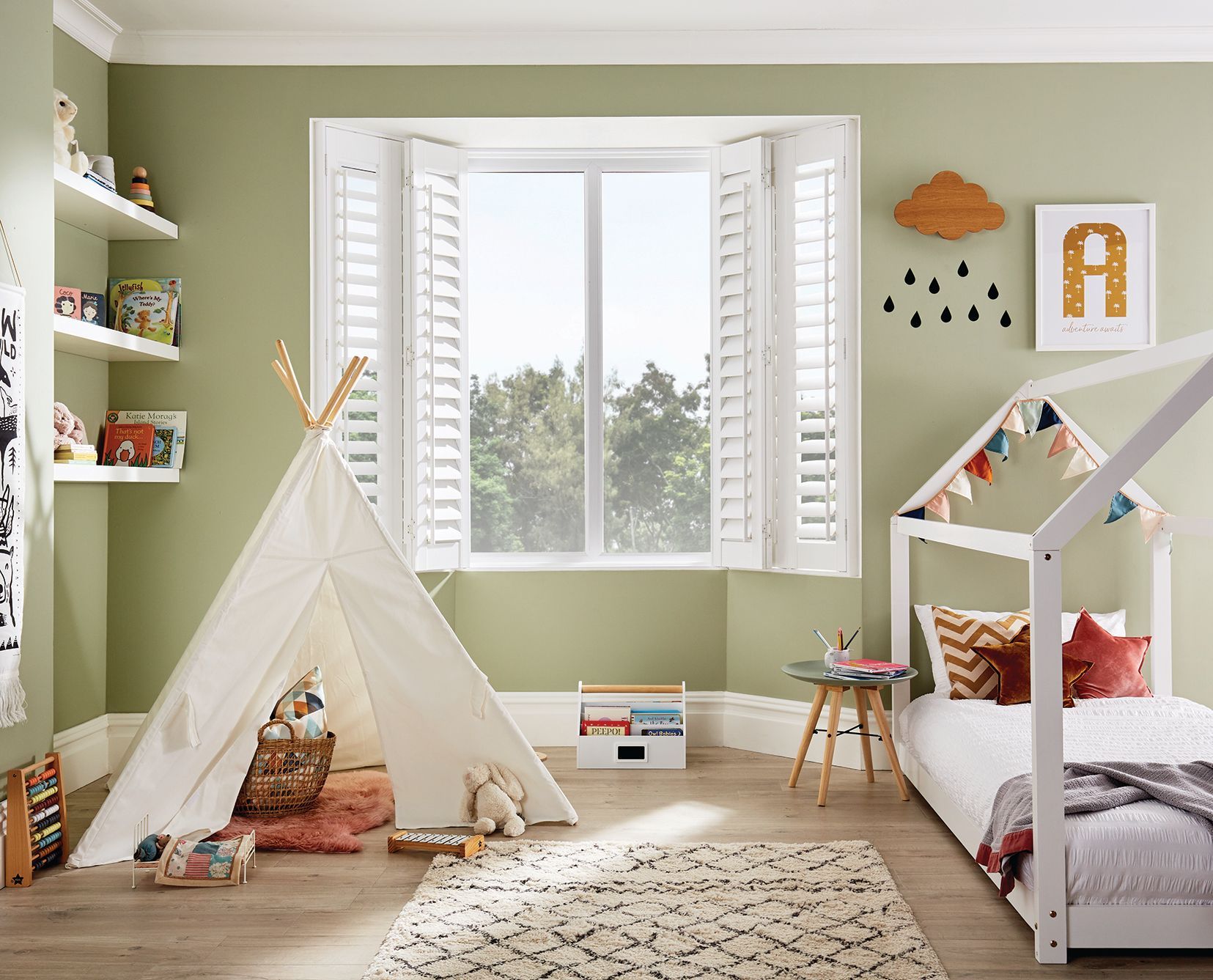 A child's bedroom with a teepee, bed, shelves, and window with white shutters; olive green walls.