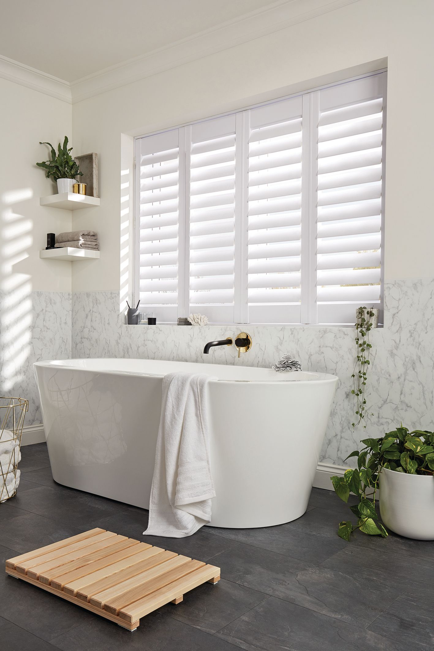 White bathroom with a bathtub, shutters, plants, and a wooden mat.