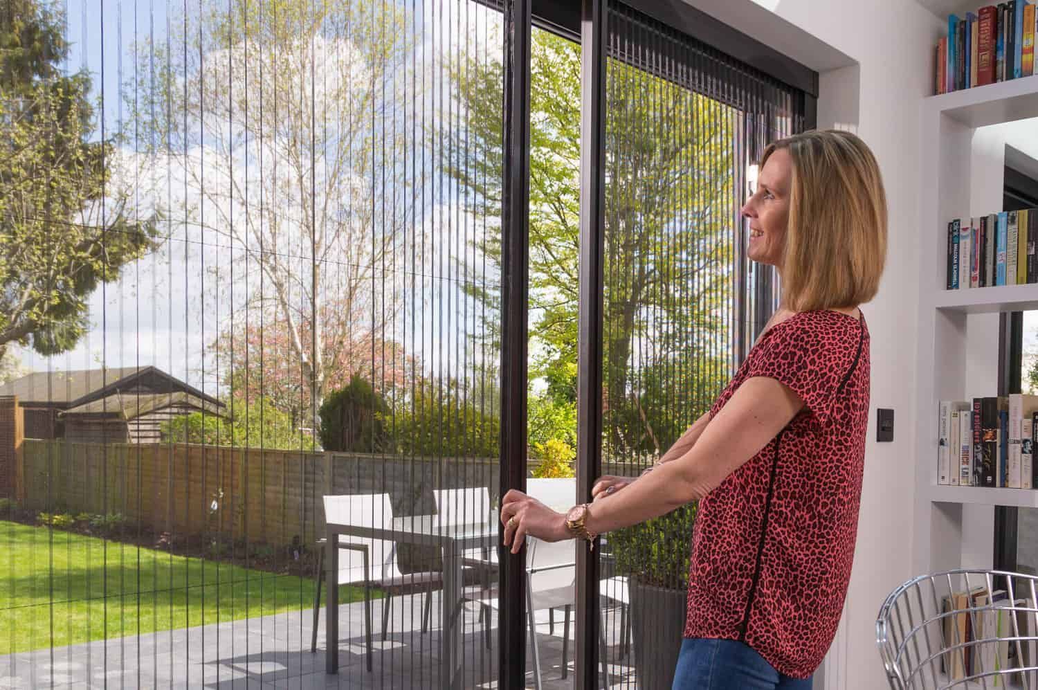 Woman opening a sliding screen door, looking out at a backyard with a table and chairs.