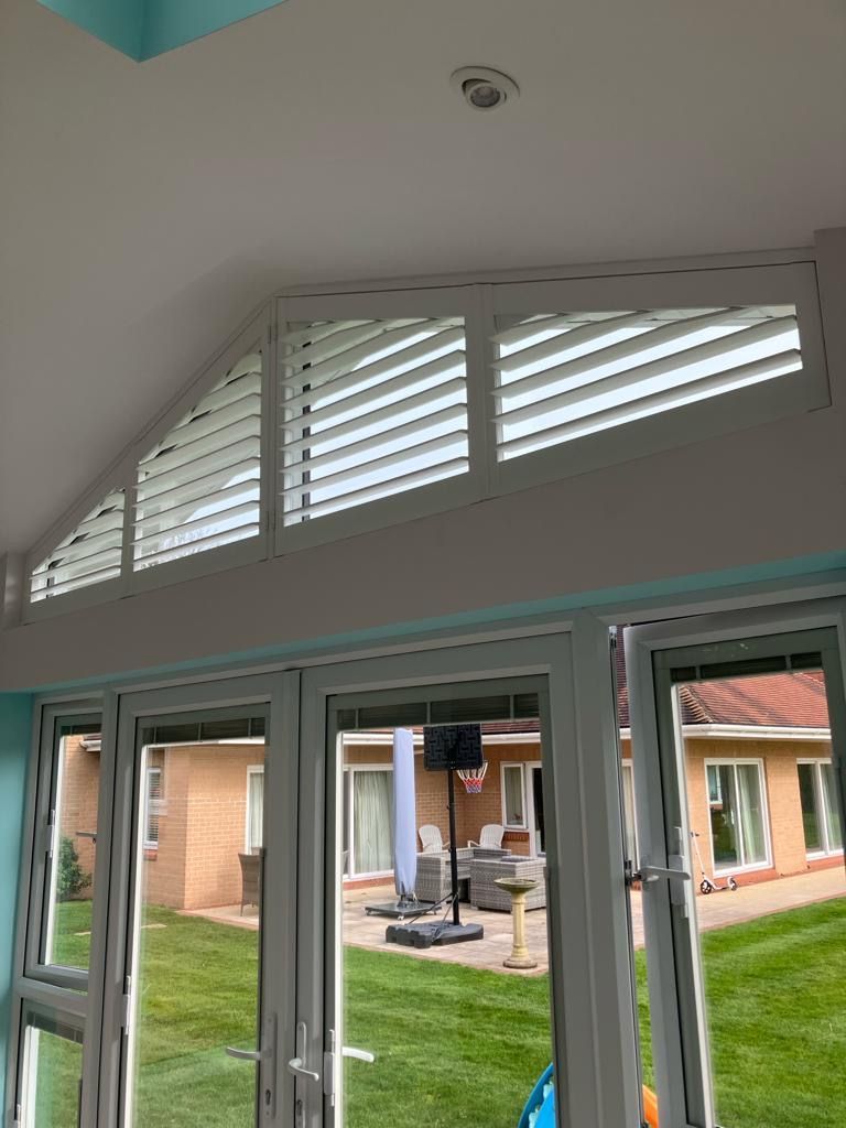 Interior view of a conservatory with white window frames and slatted vents. Green yard visible through windows.