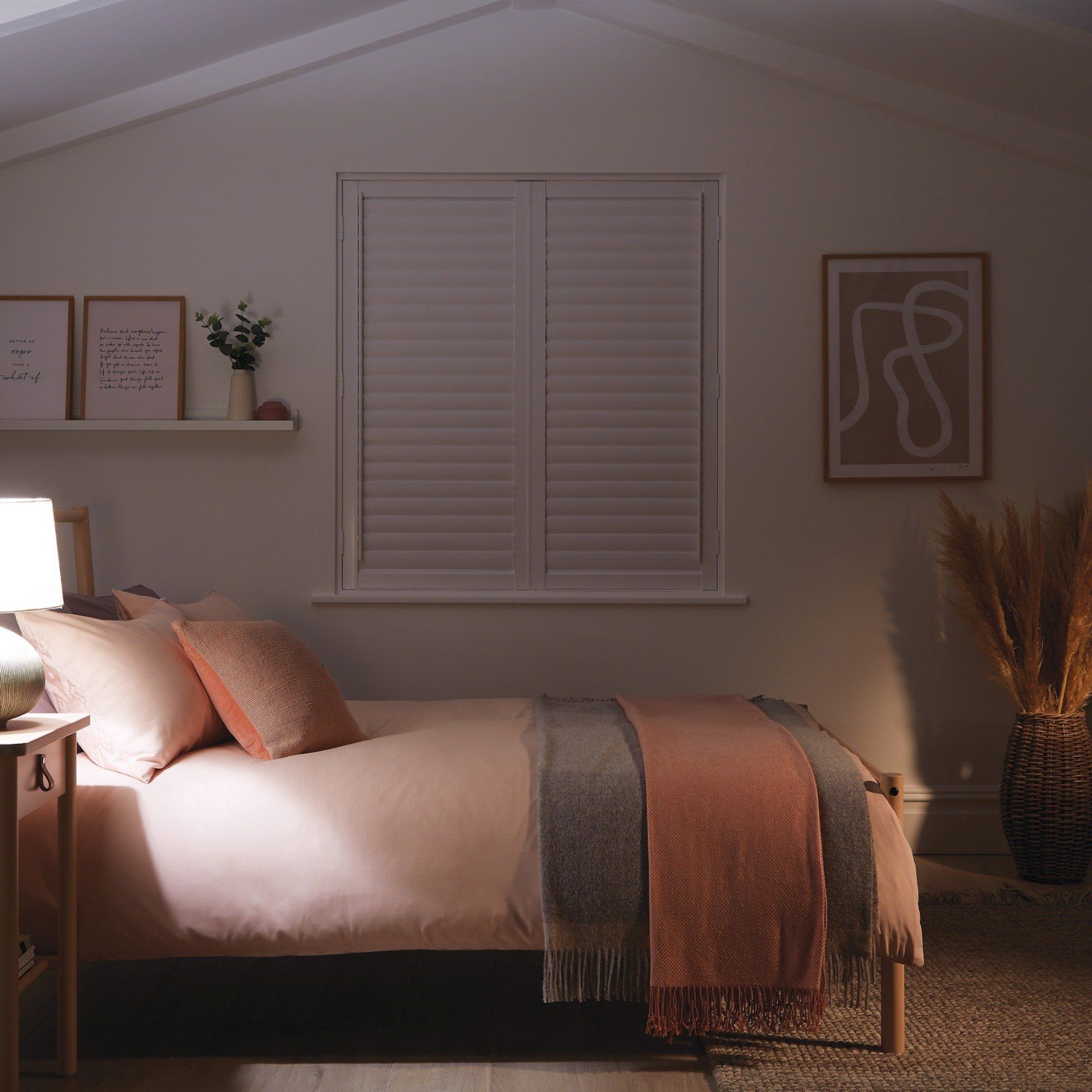 Bedroom with pink bedding, white shutters, artwork, and a bedside lamp.