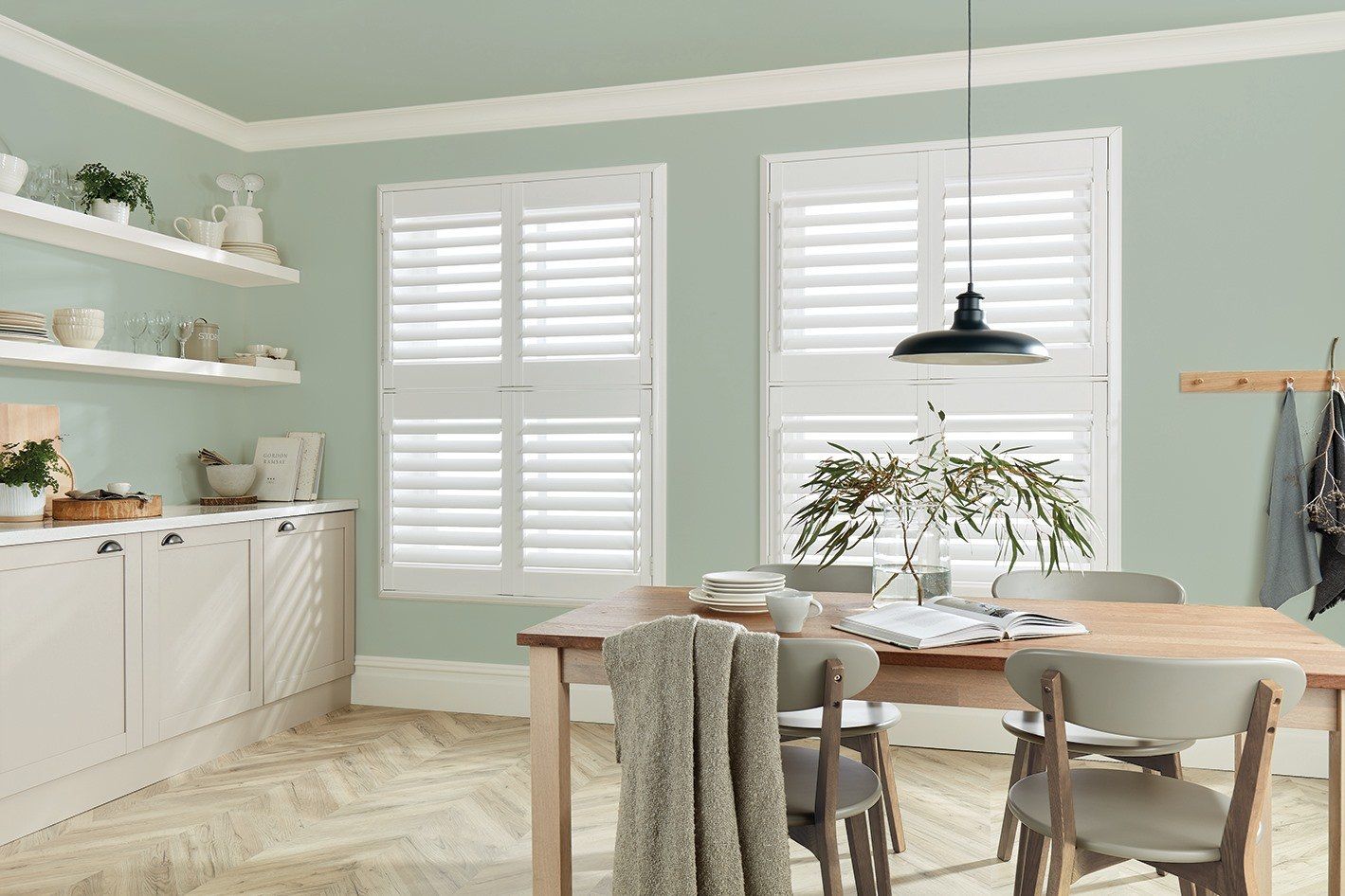 A dining room with white shutters, a wooden table, chairs, and light green walls.