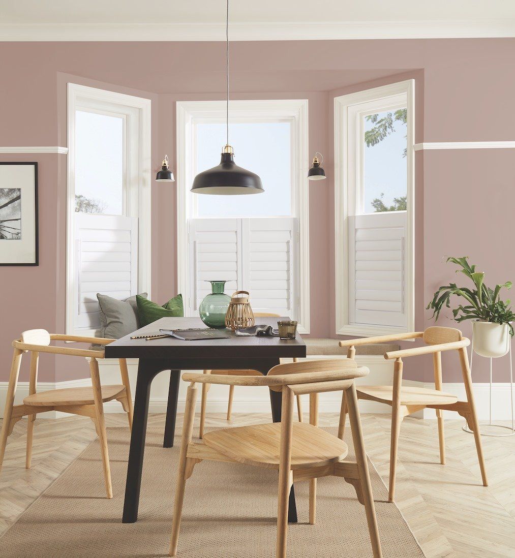Dining room with pink walls, wood chairs, black table, and windows with shutters.