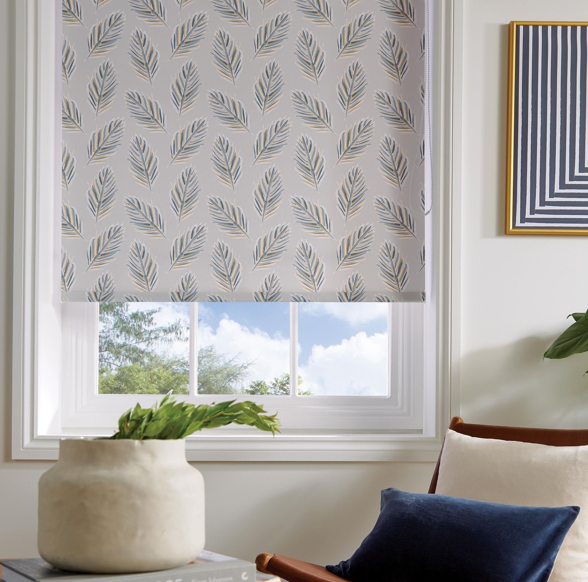 Roller blind with a leaf pattern over a window, with a vase and chair.