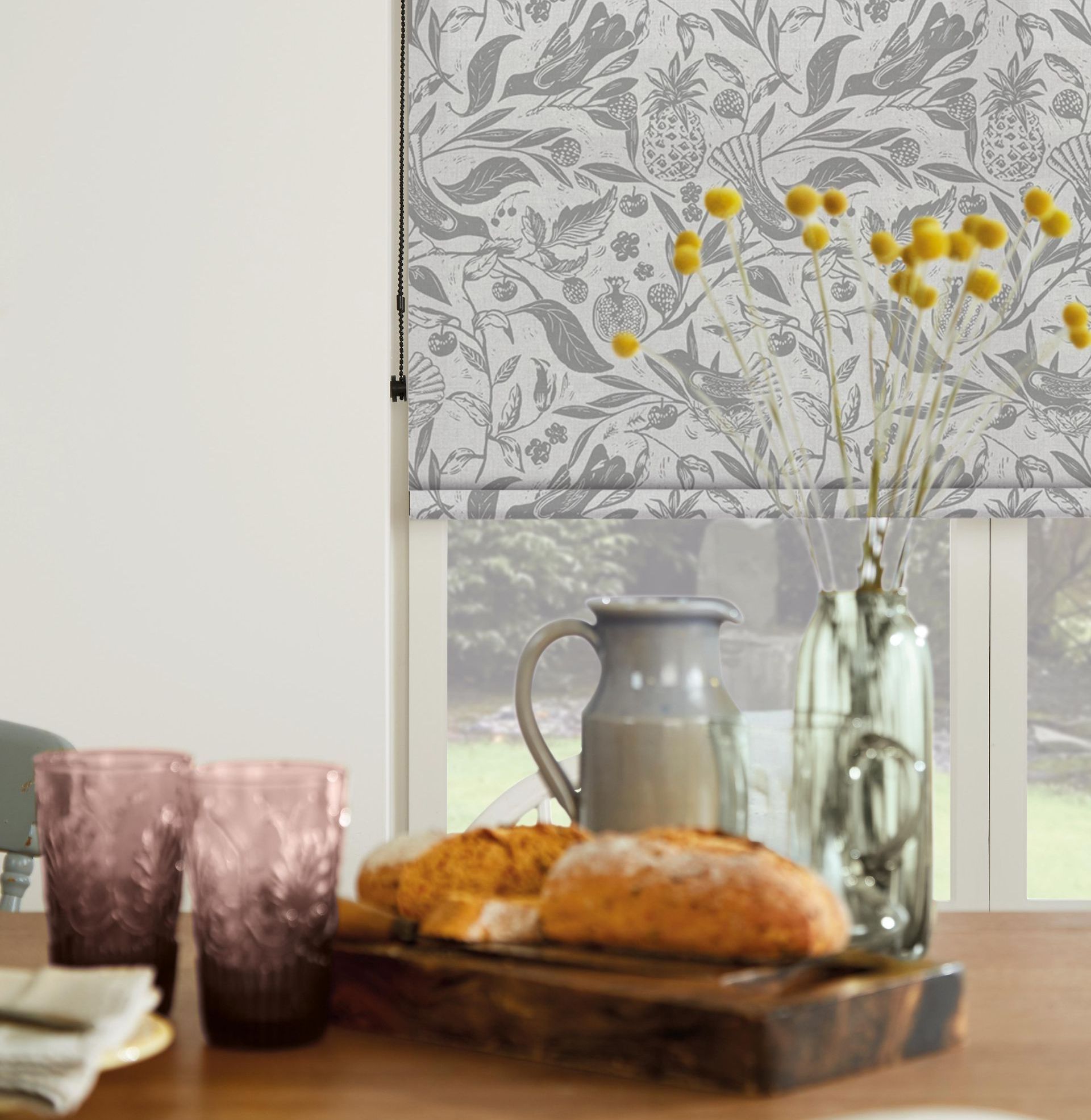 Dining table set for a meal, featuring bread, glasses, pitcher, and a floral-patterned window shade.