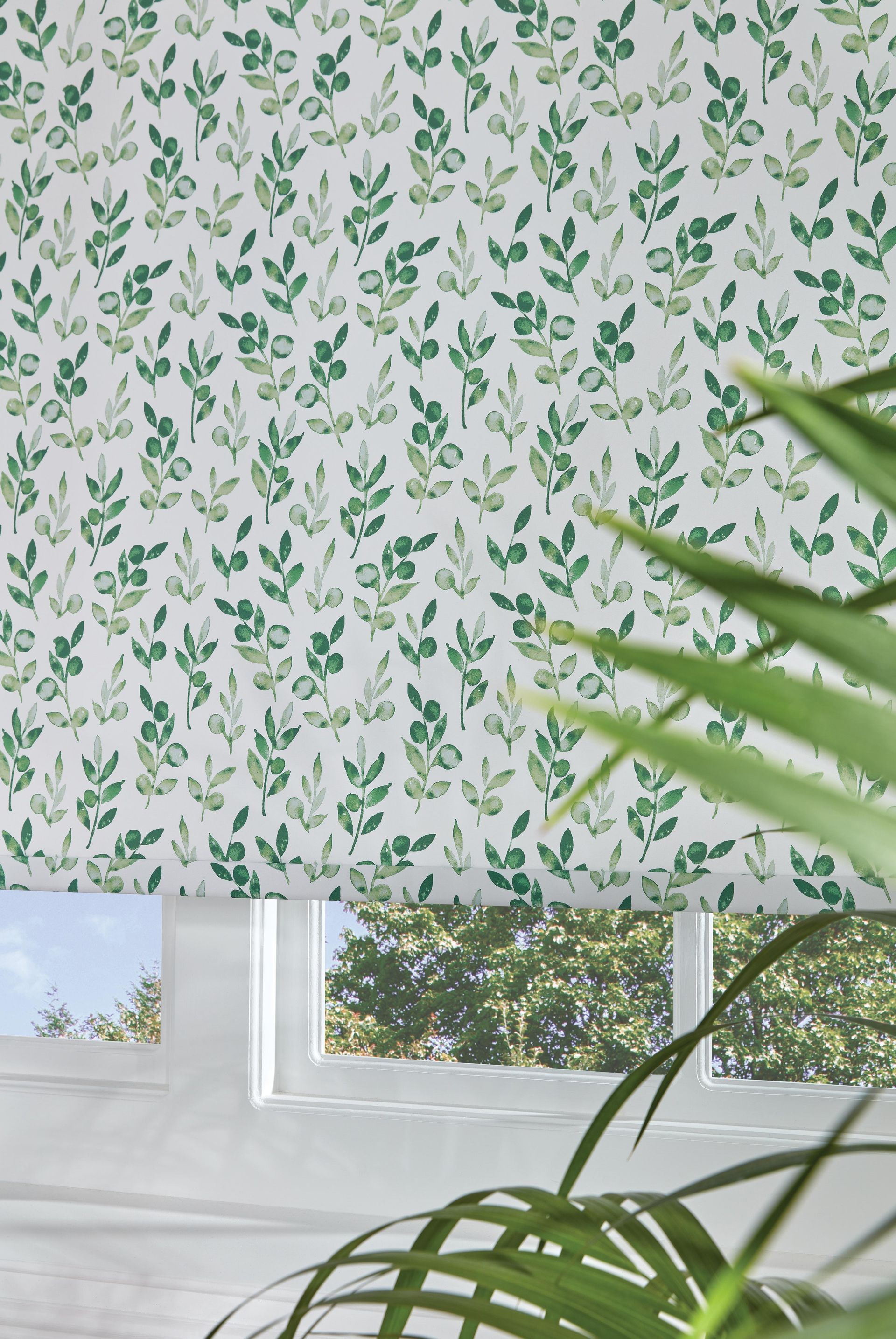White roller blind with green leaf pattern covers window. Sunny outdoor view visible.