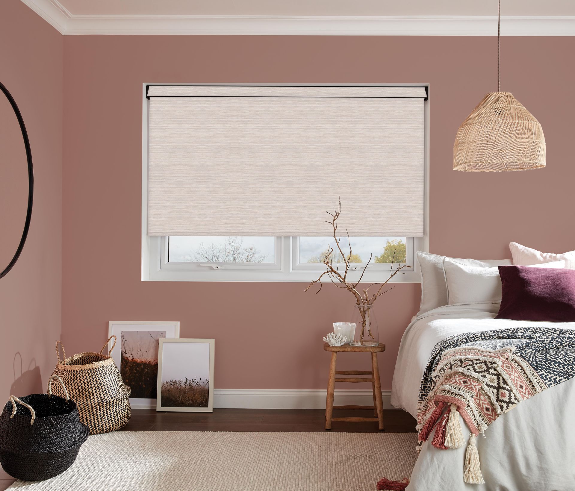 Bedroom with pink walls, window with beige roller shade, bed with pillows and blanket, and woven decor.