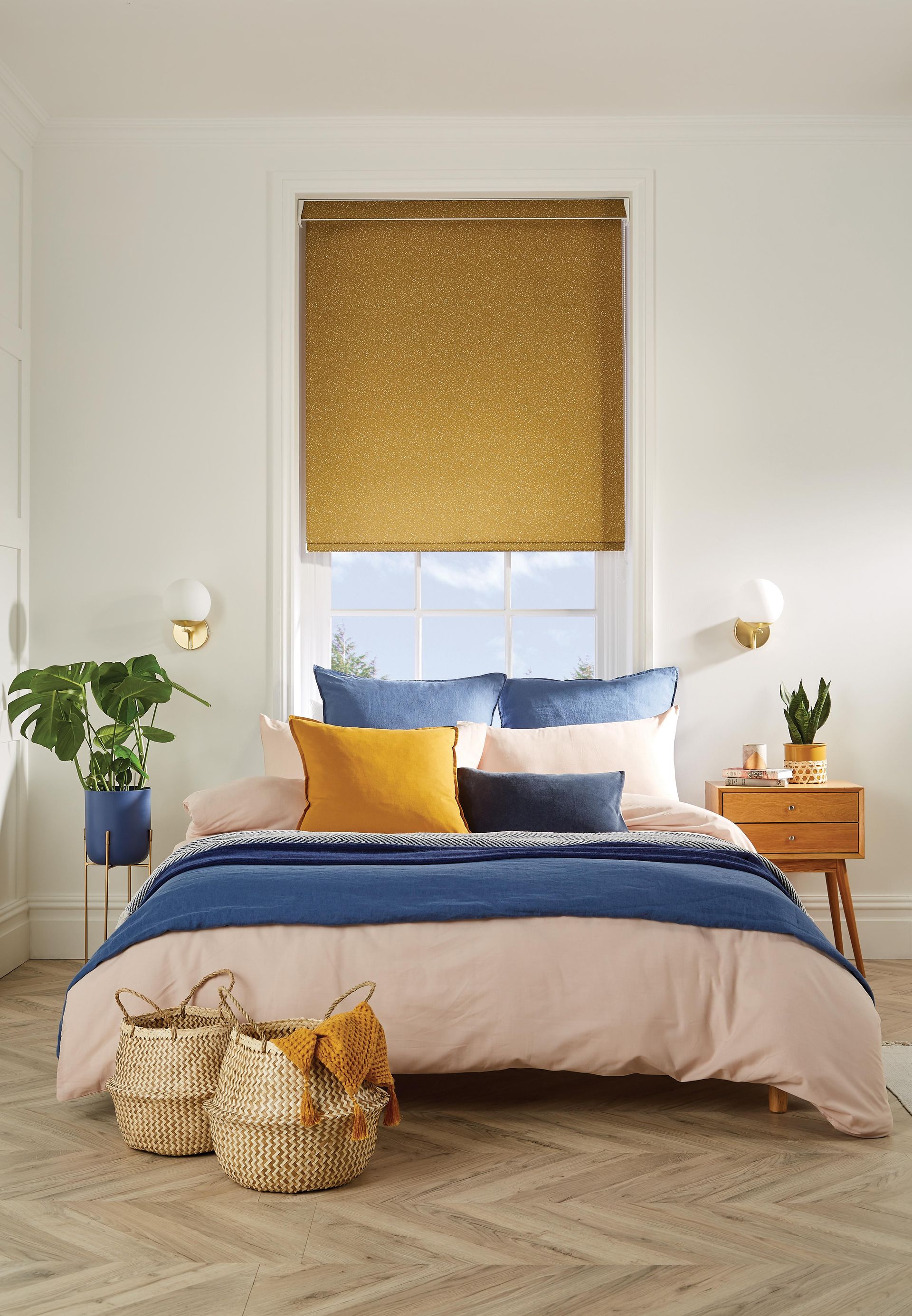 Bedroom with bed featuring pink, blue, and yellow bedding. Gold patterned window blind, plants, and side table.