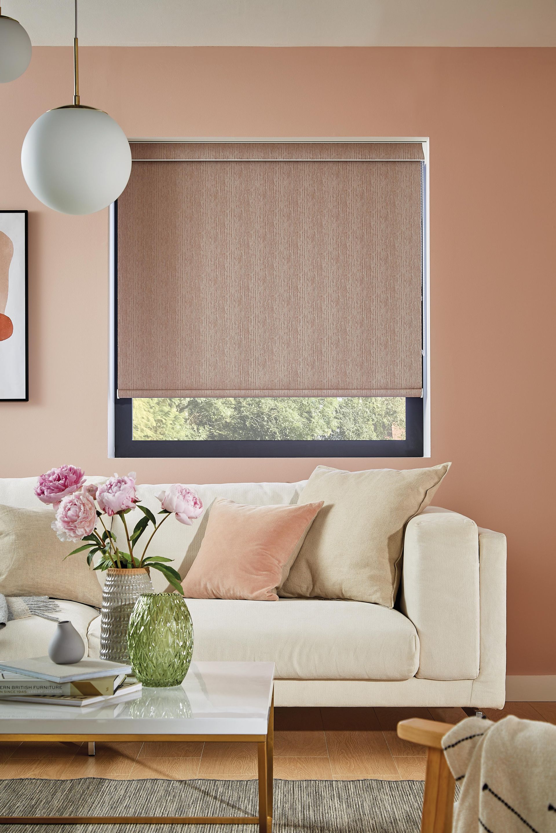 Living room with pink wall, beige couch, window with brown shade, and glass vase of flowers.